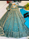 A teal blue-green Anarkali gown with gold embroidery and small white polka dots, laid flat on a white textured surface alongside a turquoise dupatta, black sandals, and decorative flowers and plants.
