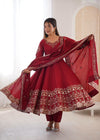 A woman modeling a deep red Indian ethnic suit with gold embroidery, featuring a long kurta, matching pants, and a dupatta, set against a bright, modern indoor background.
